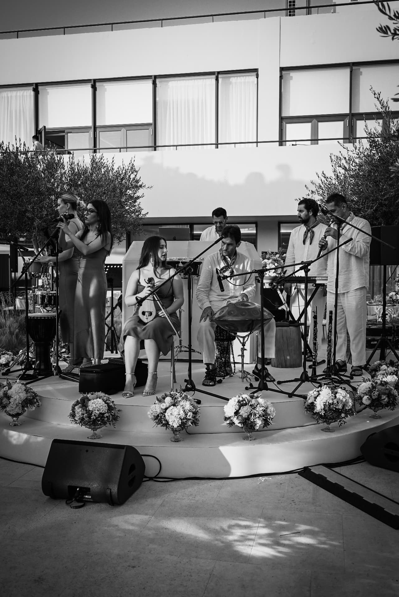 The full ensemble performing on stage in black and white, with lyra, handpan, clarinets, percussion and keyboards